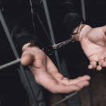 A man in handcuffs is holding onto the bars of his cage.